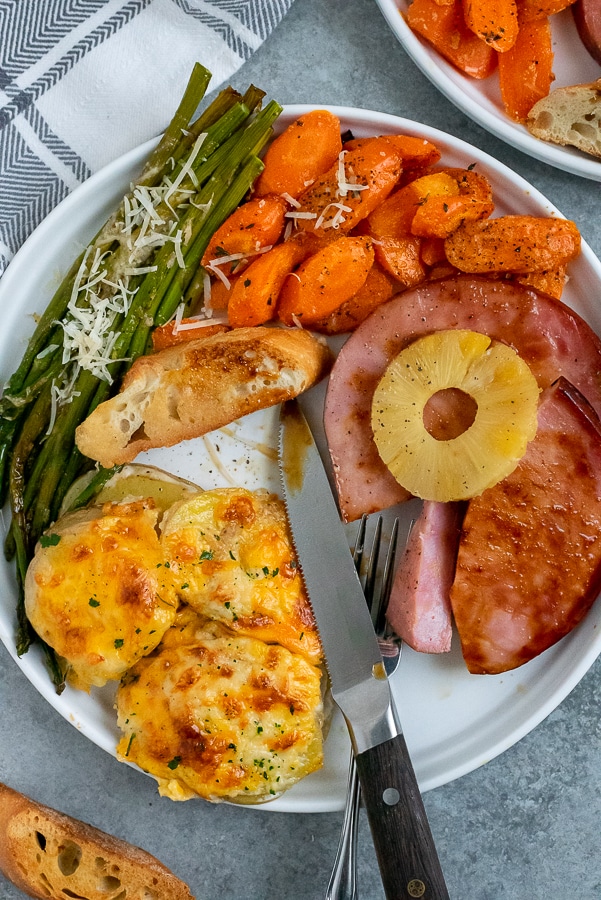 White plate filled with ham, cheesy potatoes, asaparagus, and carrots.